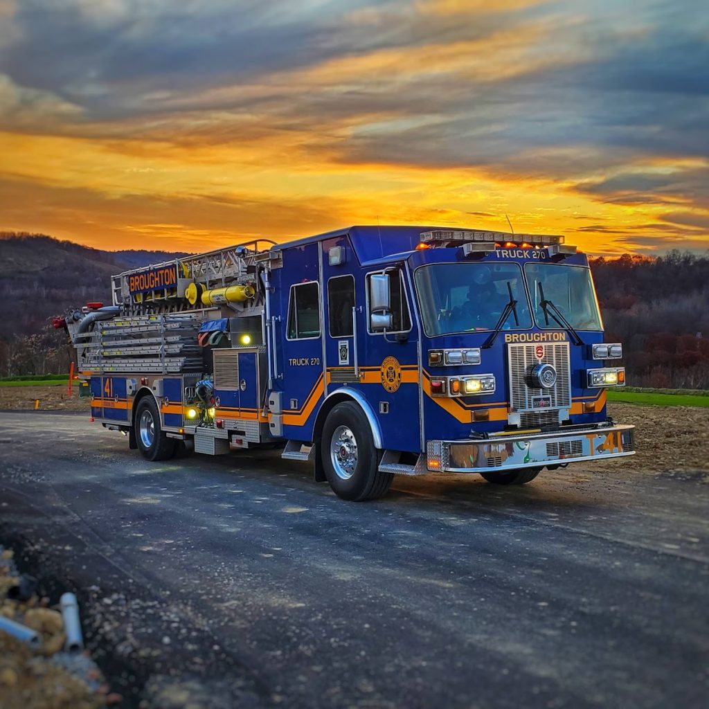 A fire truck labeled "Broughton" is parked on a road during sunset. The truck is blue and yellow with various equipment on its sides. The background features a dramatic sky with orange, yellow, and blue hues, and rolling hills.