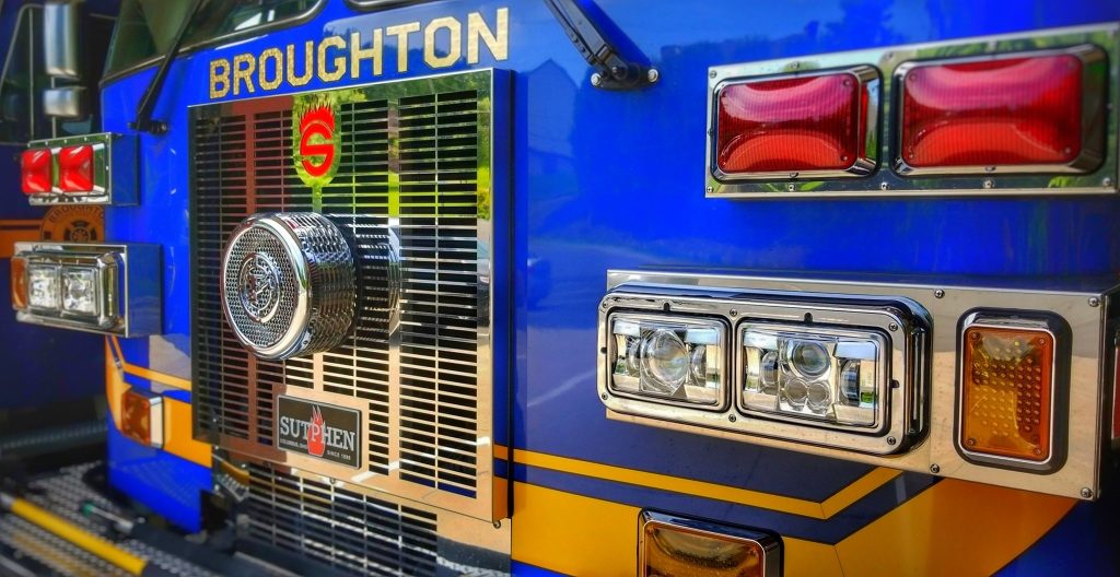 Close-up of the front of a blue fire truck with the word "BROUGHTON" displayed at the top. The front also features shiny chrome details, including a prominent circular grille, and various lights in red and clear colors.