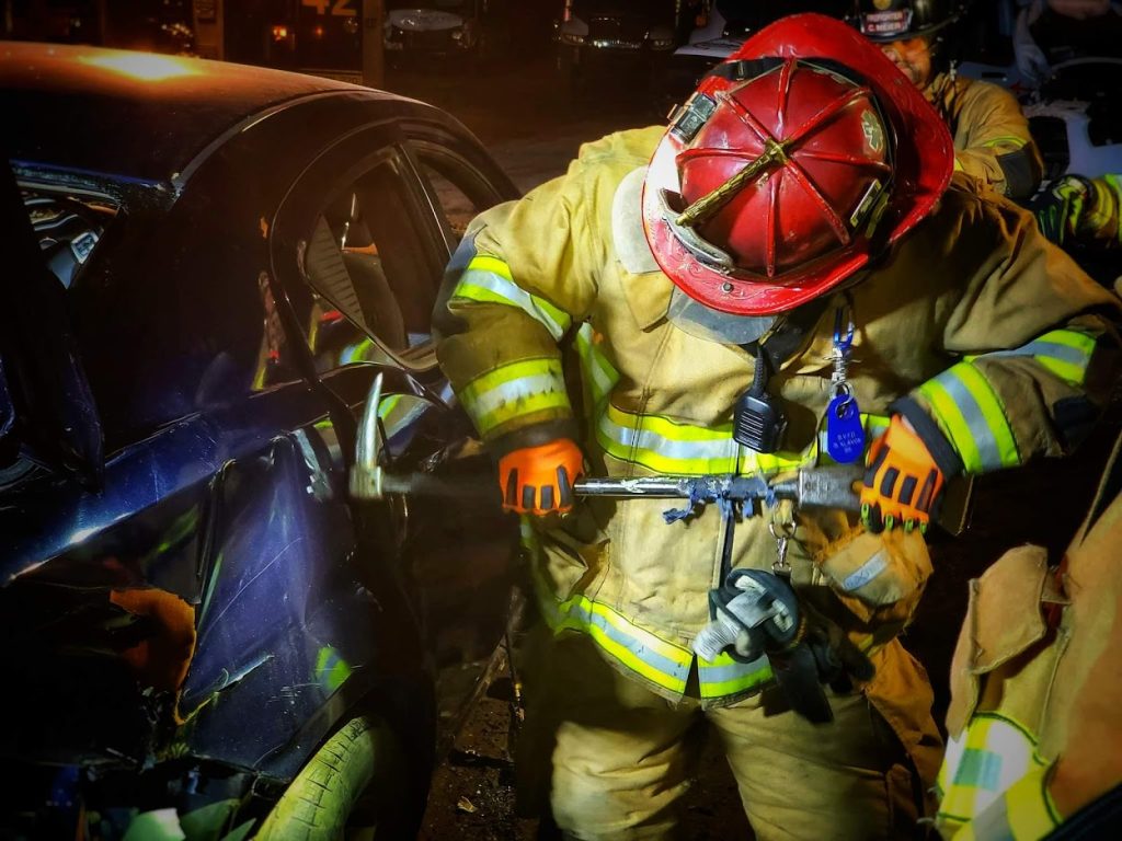 A firefighter in full gear, including a red helmet, uses a specialized tool to extricate someone from a damaged vehicle at night. The vehicle's door appears severely dented, and another firefighter is partly visible in the background.