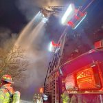 Firefighters work to extinguish a fire at night, using a ladder truck to spray water over a building. The image shows three firefighters in full gear with helmets observing the scene, while smoke billows around them and the truck's emergency lights flash.