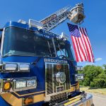 A bright blue fire truck with "Broughton" written on it extends a ladder holding a large American flag. The scene is set outdoors on a sunny day with a clear blue sky and green trees in the background.