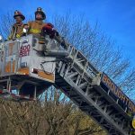 Two firefighters in full gear, including helmets and protective clothing, stand atop an extended fire truck ladder labeled "BROUGHTON" and "500 FEET". They are surrounded by leafless trees against a clear blue sky.