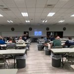 A group of people sits scattered across a large room with tables and chairs, facing a presentation screen. Two presenters stand at the front, and a slideshow is displayed on the screen. Trash bins are placed along the rows of tables.