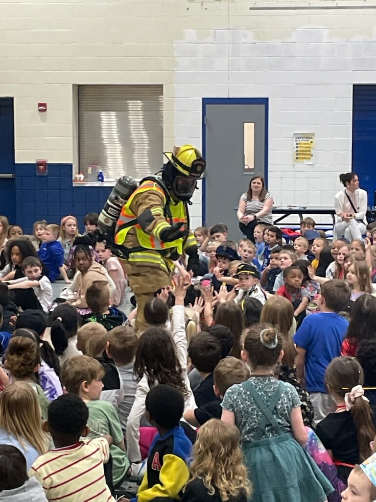 A firefighter in full gear, including helmet and oxygen tank, interacts with a large group of schoolchildren seated on the floor in what looks like a gymnasium. The children eagerly raise their hands, and a few adults are sitting in the background.