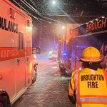 Emergency responders including ambulance and fire truck teams, labeled "TRI-COMMUNITY EMS" and "BROUGHTON FIRE," are on a snow-covered street at night. Bright lights illuminate the scene as responders assess the situation. Snow falls thickly, adding to the urgency.