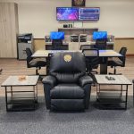 A modern office space features three black leather recliners with embroidered lion logos arranged in front of two small tables. Behind them, a long conference table with two computer monitors on stands, files, and a printer. In the background are a kitchen area and multiple screens displaying sports.
