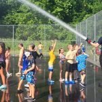 A group of children is joyfully playing on a wet basketball court as two firefighters spray water from a hose. The children are dressed in swimwear, running around and splashing in the water. The scene is lively, with green trees in the background and a bright, sunny sky.