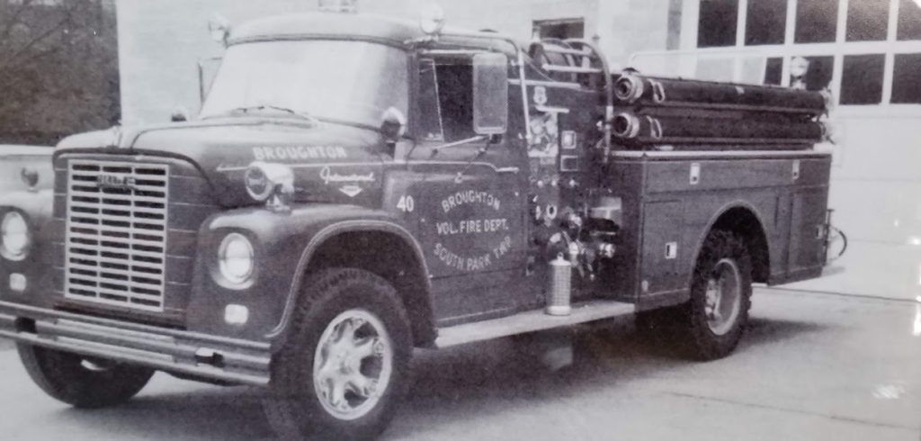 A vintage Broughton Volunteer Fire Department fire truck is parked outside a fire station. The truck is equipped with ladders, hoses, and various firefighting equipment. It has "Broughton Vol. Fire Dept." and the number "40" written on the side.
