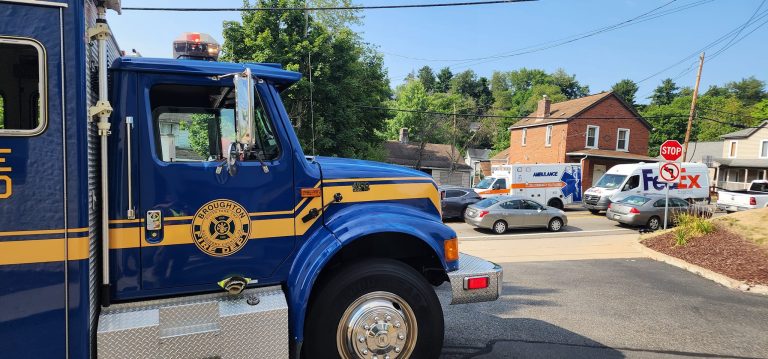 A blue fire truck is parked on a suburban street near a stop sign. In the background, there is a white ambulance and multiple parked cars. Houses with trees are visible in the distance under a clear blue sky.