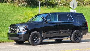 A black SUV is parked on the side of a road during the day. The vehicle has dark-tinted windows and off-road tires. The words "COMMAND 210" are printed on its side. In the background, there is green grass and some bushes.