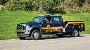 A dark blue fire rescue truck with "Broughton" written on the side, parked on the side of a road. The truck is equipped with various emergency response tools and has a yellow stripe along the side. In the background is a grassy area with trees.