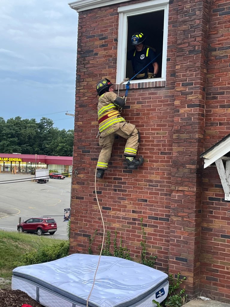 A firefighter in full gear climbs up a brick building using a rope, while another firefighter assists from a second-story window. A mattress is placed on the ground below for safety. A Dollar General store and parking lot are visible in the background.