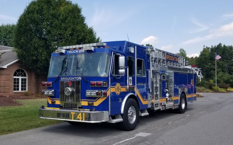 A blue and yellow fire truck labeled "Broughton" and "Truck 270" is parked on a residential street. The truck has an extended ladder on top. A house and trees are in the background, with an American flag on a pole visible to the right.