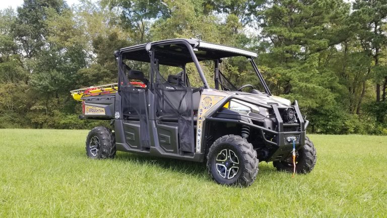 An all-terrain utility vehicle with emergency rescue equipment mounted on the rear is parked on a grassy field. The vehicle has four seats, a winch on the front bumper, and is marked with "Station 2-81" on the side. Trees are visible in the background.