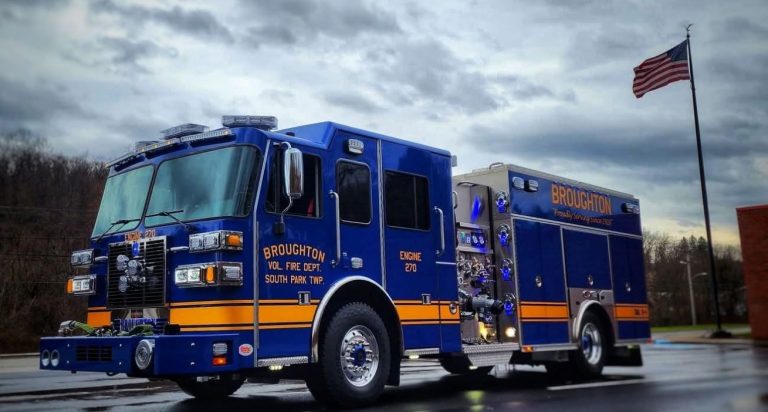 A blue and yellow fire truck labeled "Broughton Vol. Fire Dept. South Park Twp, Engine 270" is parked on a street. An American flag is flying in the background with a cloudy sky above.