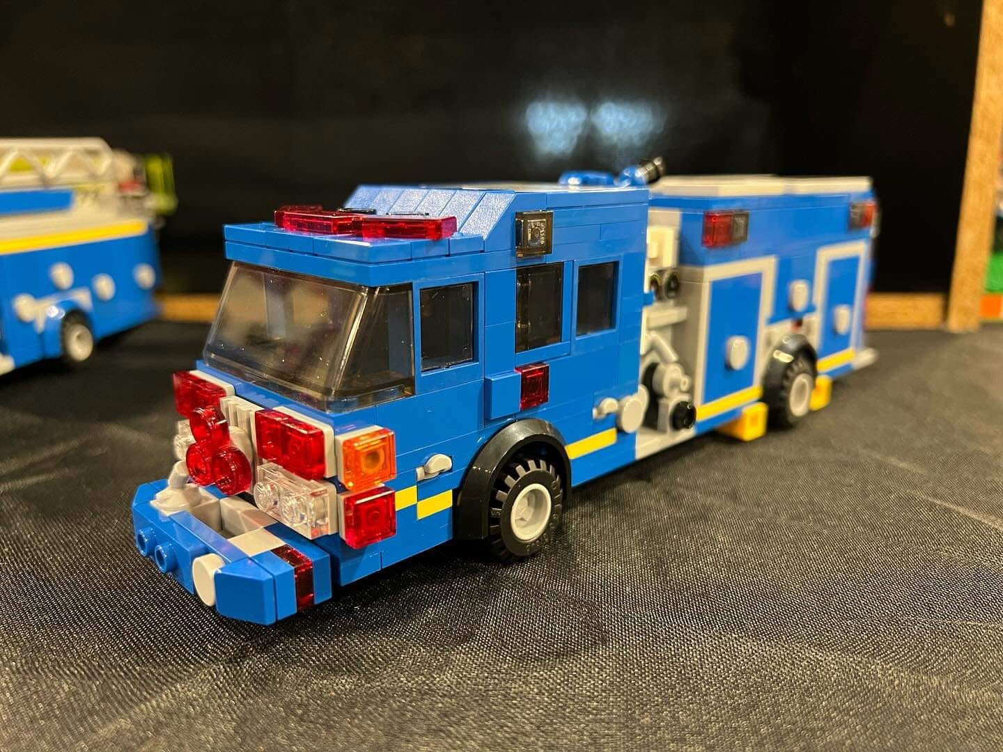 A detailed Lego fire truck model with a blue and yellow color scheme. The truck features transparent windows, red lights on the roof, front, and sides, and various accessories on its sides and rear. The design includes intricate details and realistic elements.