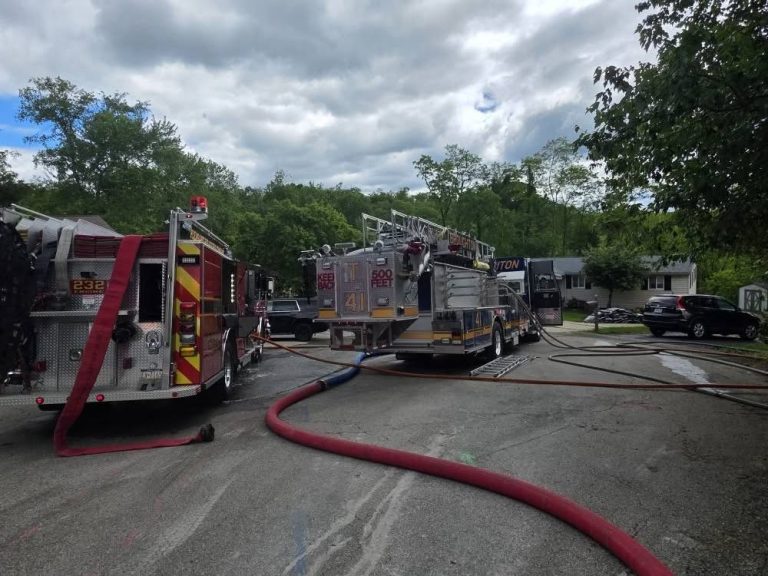 Two fire trucks with extended hoses are parked on a residential street with trees and houses in the background. Firefighters and equipment are visible near the trucks under a cloudy sky.