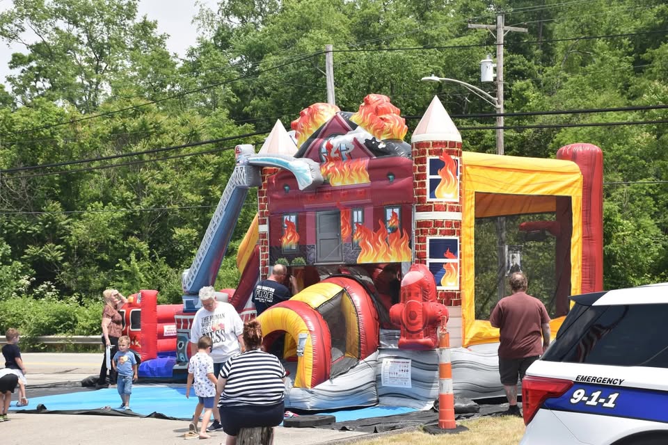 A fire truck-themed inflatable bounce house with kids and adults nearby at an outdoor event. A police car labeled “Emergency 9-1-1” is parked on the right. Trees and power lines are in the background.